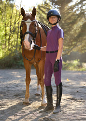 Full body front view of a young female model wearing the purple Kerrits Kids Coolcore Full Seat Tech Tights and coordinating purple shirt while standing in front of a chestnut horse