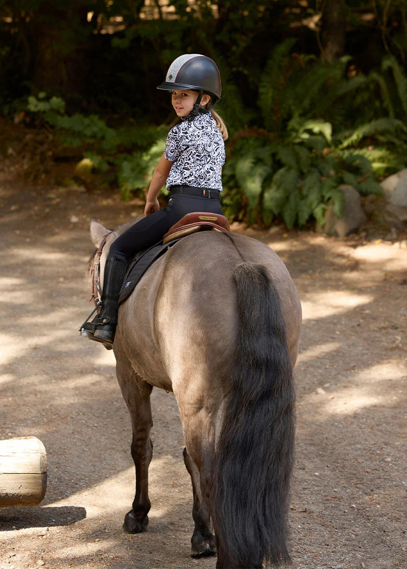 Back view of a young female model wearing the black Kerrits Kids Coolcore Full Seat Tech Tights while riding a horse outside
