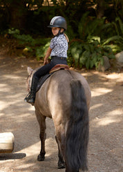 Back view of a young female model wearing the black Kerrits Kids Coolcore Full Seat Tech Tights while riding a horse outside