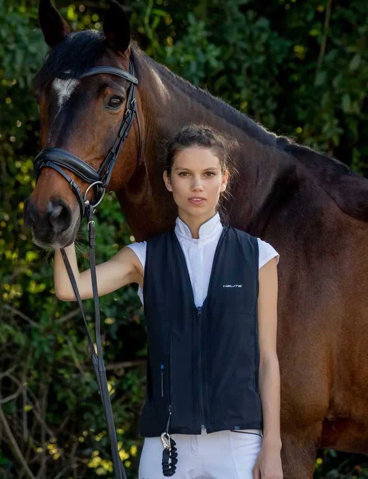 Model wearin the Helite Zip'In 2 Airbag in black with white breeches standing next to a bay horse