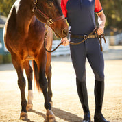 Front view of a male model wearing the Navy Ariat Men's Tri Factor Knee Patch Breeches while leading a horse on a path outside