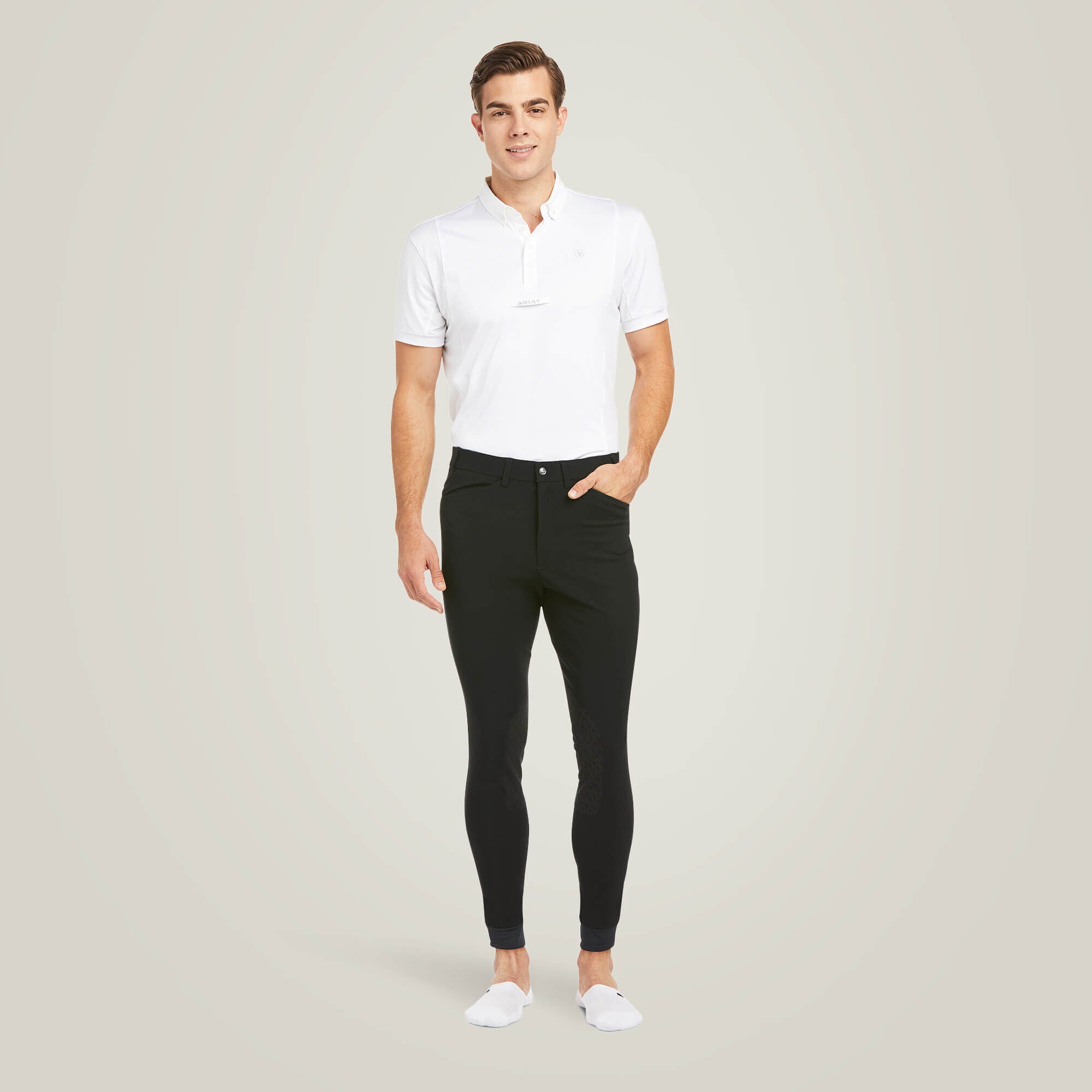 Full body front view of a male model wearing the Black Ariat Men's Tri Factor Knee Patch Breeches with a white shirt on a plain background