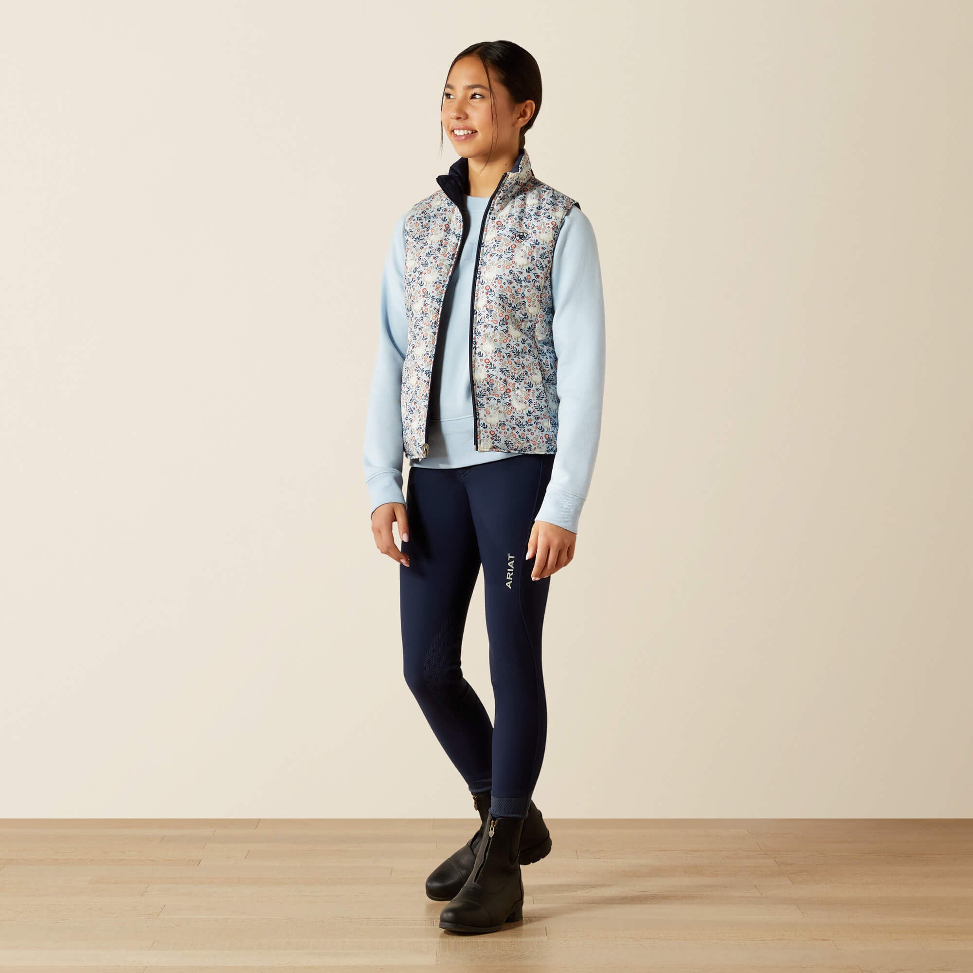 Full body front view of a young female model wearing the Posey Pony Print Ariat Youth Bella Reversible Insulated Vest with blue breeches on a plain background