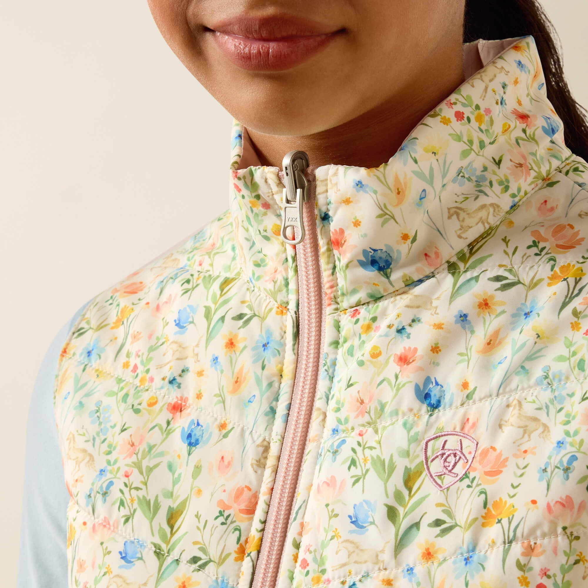 Close-up front view of a young female model wearing the Horse In Flowers/Sepia Rose Ariat Youth Bella Reversible Insulated Vest on a plain background