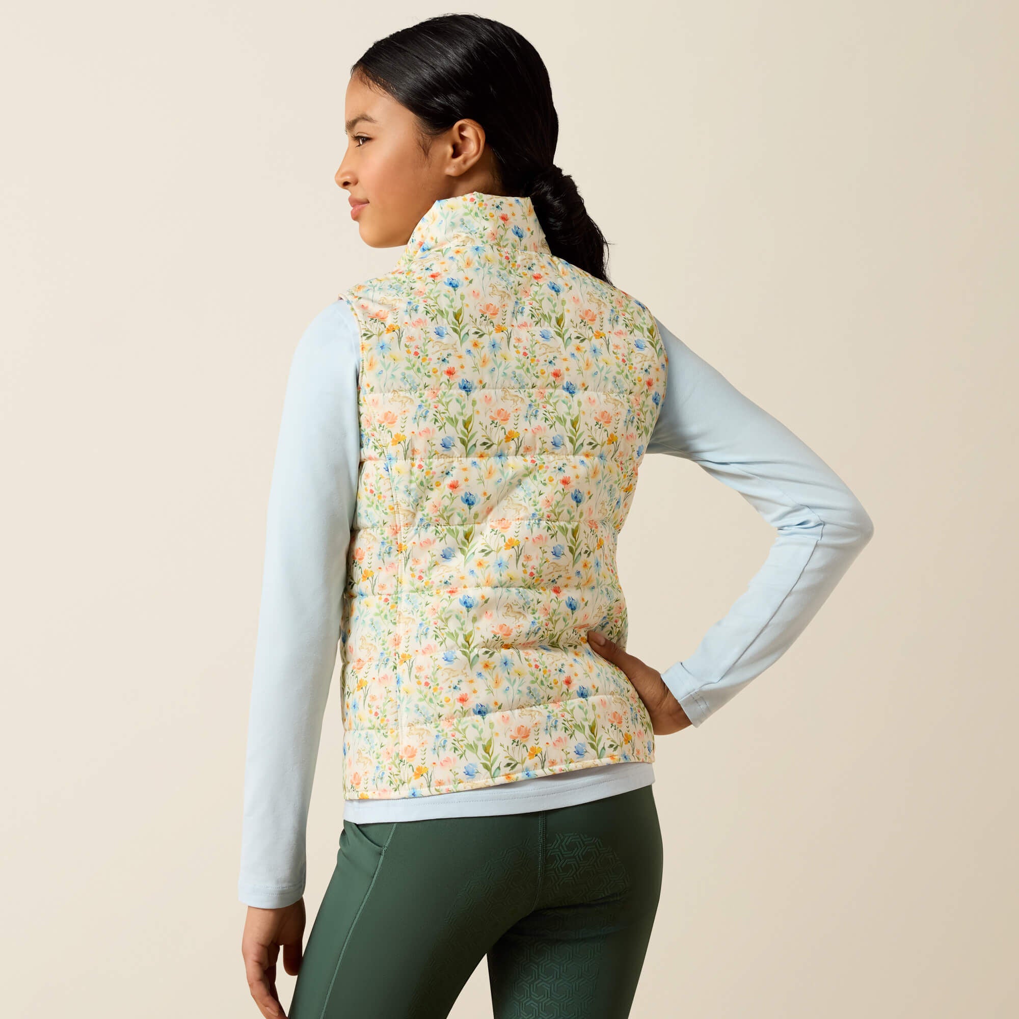 Back view of a young female model wearing the Horse In Flowers/Sepia Rose Ariat Youth Bella Reversible Insulated Vest on a plain background
