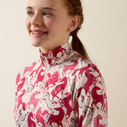 Angled front closeup view of a young female model wearing the Vanilla Horsin' Around Print Ariat Youth Lunaveil 1/4 Zip Long Sleeve Baselayer on a plain background showing the upper half of the baselayer