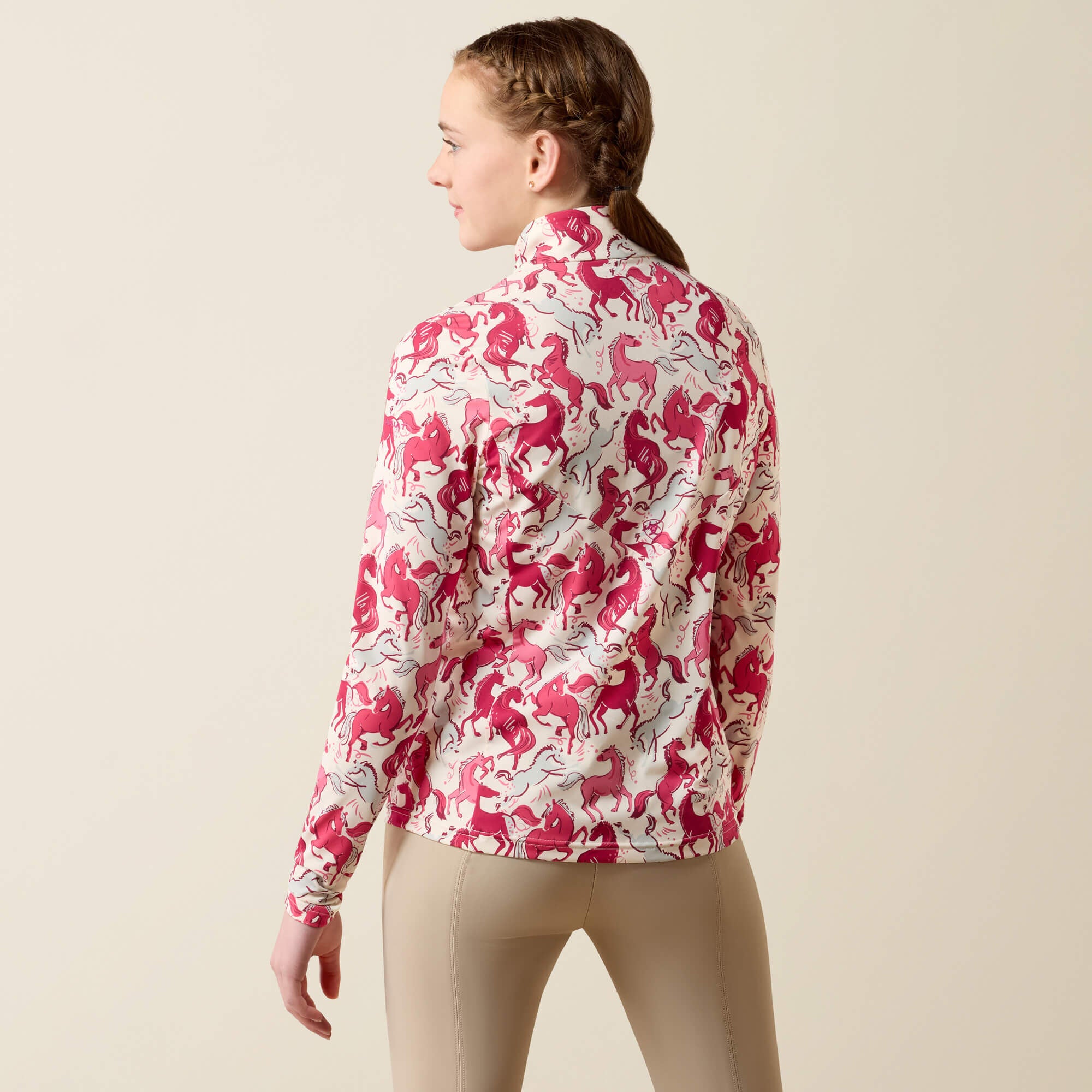 Back view of a young female model wearing the Vanilla Horsin' Around Print Ariat Youth Lunaveil 1/4 Zip Long Sleeve Baselayer on a plain background