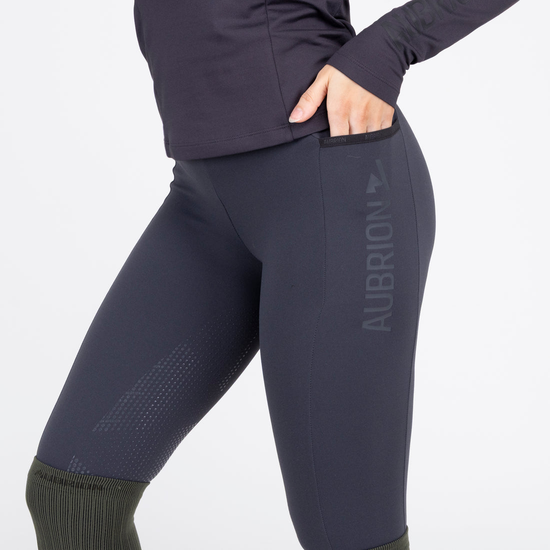 Side view of a female model wearing the Charcoal Aubrion React Winter Knee Patch Riding Tights on a plain background