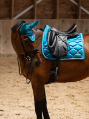 Side view a bay horse wearing the Evening Teal PS of Sweden Signature Dressage Pad while standing in an indoor arena