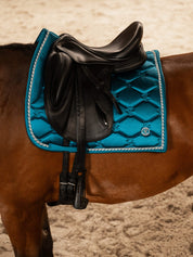 Side view of a bay horse wearing the Evening Teal PS of Sweden Signature Dressage Pad while standing in an indoor arena
