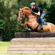 Side view of a male model riding a chestnut horse over a cross country jump while the horse is wearing the Kavalkade Jerome Breastplate