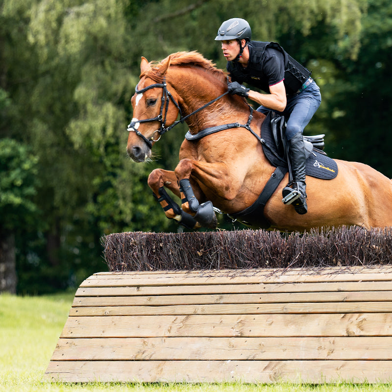 Side view of a male model riding a chestnut horse over a cross country jump while the horse is wearing the Kavalkade Jerome Breastplate