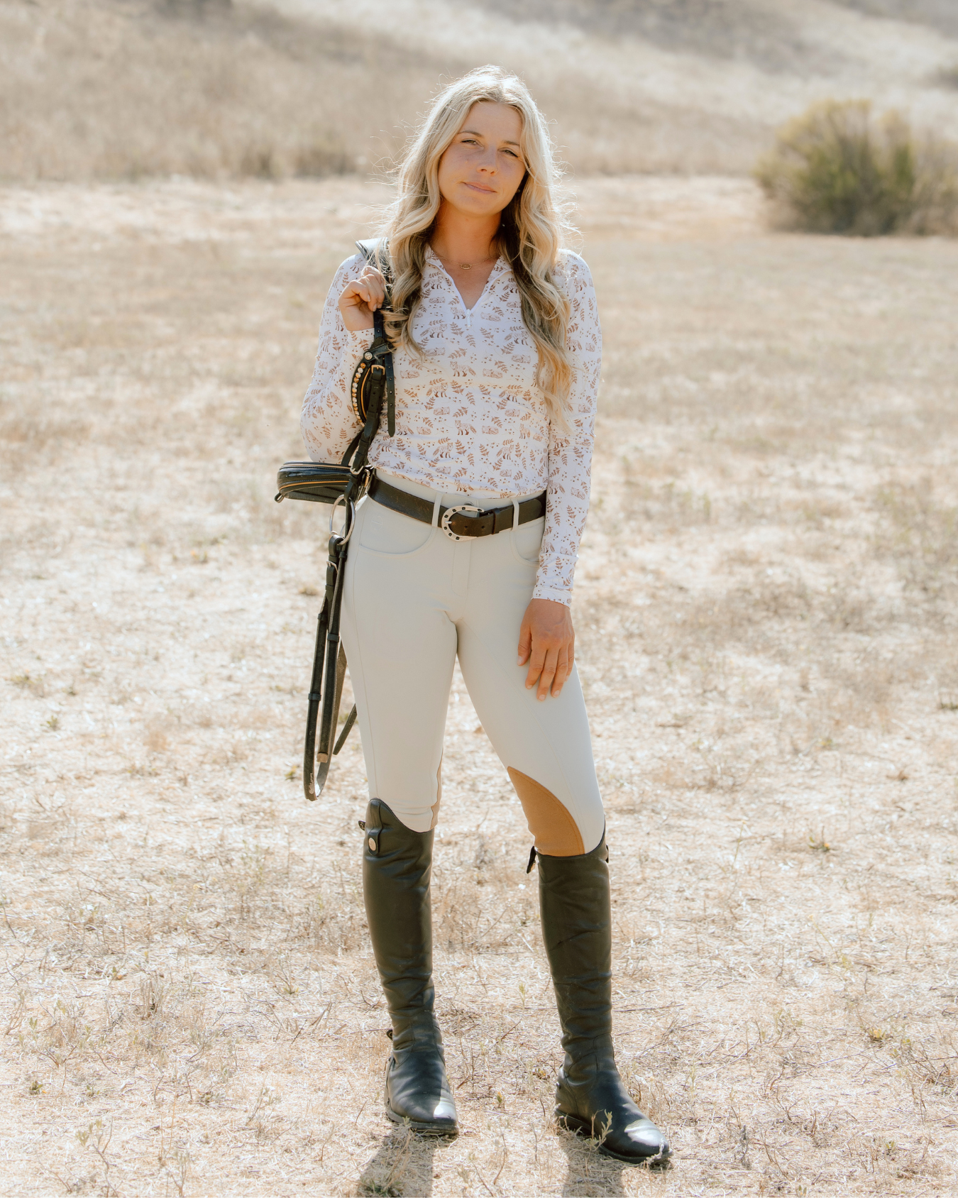 Full body front view of a female model wearing the Deinhardt Designs Impulsion & Straightness Lori Long Sleeve Signature Sunshirt and tan breeches while standing outside