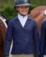 Front view of a young female model wearing the Navy Kerrits Kids Stretch Competitor Show Coat  while standing between 2 horses