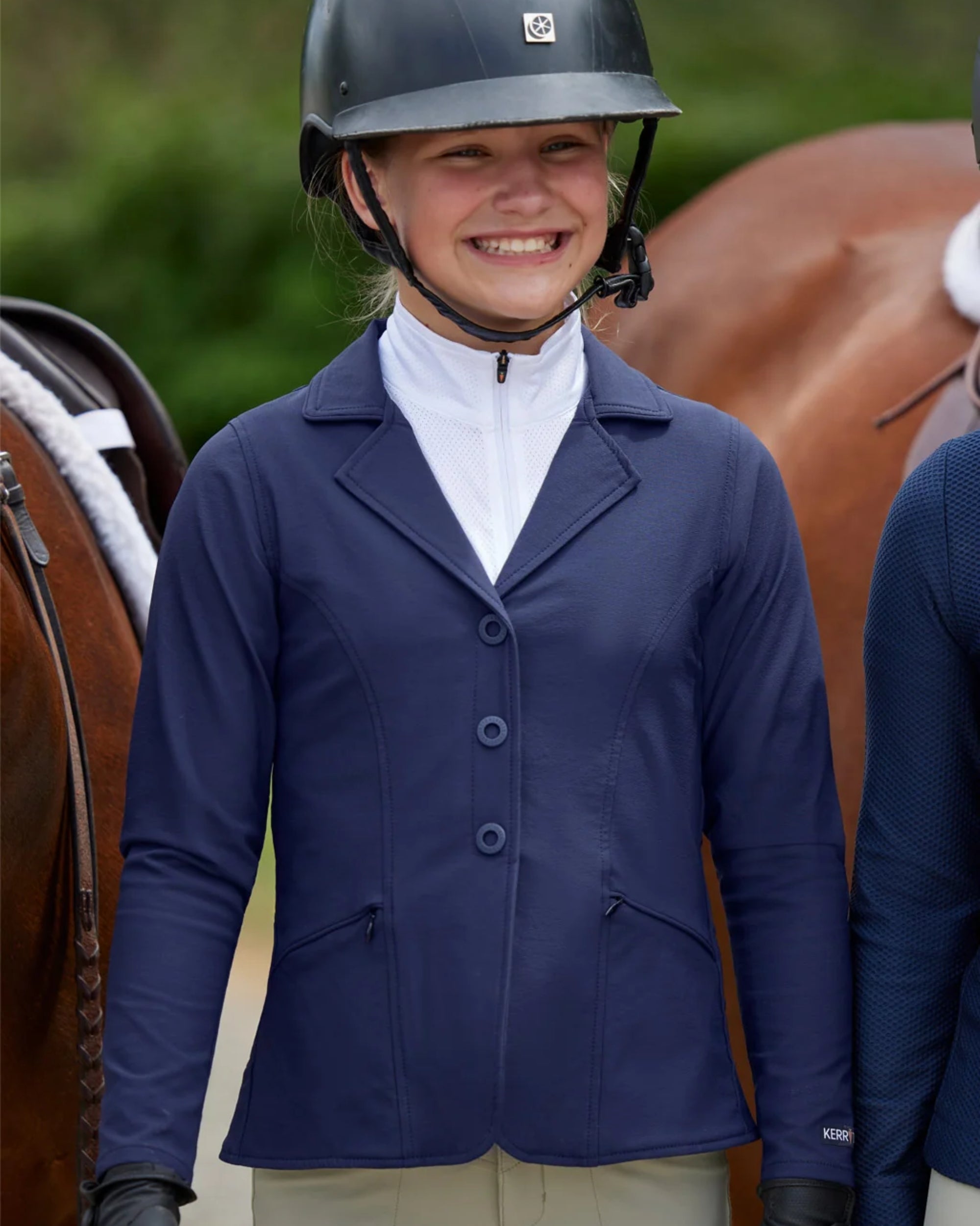 Front view of a young female model wearing the Navy Kerrits Kids Stretch Competitor Show Coat  while standing between 2 horses