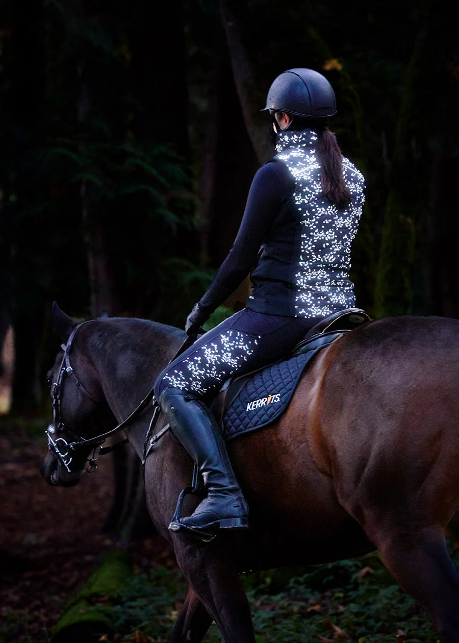 Front view of a model wearing the Black Happy Herds Kerrits Horse Reflection Quilted Vest while riding a horse on a trail in the woods