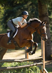 Side view of woman rider wearing the light blue Kerrits Precision Short Sleeve Quarter Zip Tech Top and gray breeches while jumping a bay horse