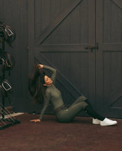 Side view of a female model wearing the Hunter Green Criniere Juliette High Compression Half Seat Breeches while sitting on the ground in front of a barn