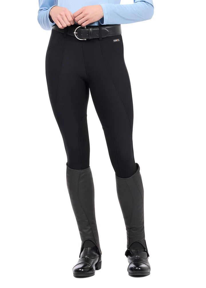 Front view of model wearing the Kerrits Microcord Knee Patch Pocket Tight in black with half chaps on a white background