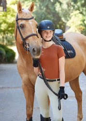 Front view of a young female model wearing the tan Kerrits Kids Coolcore Full Seat Tech Tights and orange shirt while standing in front of a palomino horse