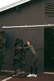 Side view of a female model wearing the Hunter Green Criniere Juliette High Compression Half Seat Breeches while leaning up against a saddle rack in front of a barn