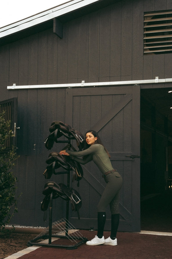 Side view of a female model wearing the Hunter Green Criniere Juliette High Compression Half Seat Breeches while leaning up against a saddle rack in front of a barn