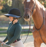 Side view of a female model wearing the black EquiVisor Sport Sun Visor while standing in front of a horse in an arena