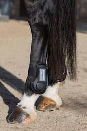 Close-up side view of a black horse wearing a pair of black Professional's Choice Pro Performance Open Rear Boots while standing in sand