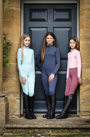 View of 3 young female models standing in front of a door while wearing coordinated LeMieux outfits, with the middle model wearing the Dusk Blue LeMieux Young Rider Lizzie Mesh Legging with matching shirt
