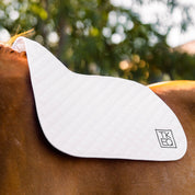 Angled side view of a horse wearing the White TKEQ x SPR Jump Saddle Pad while standing outside