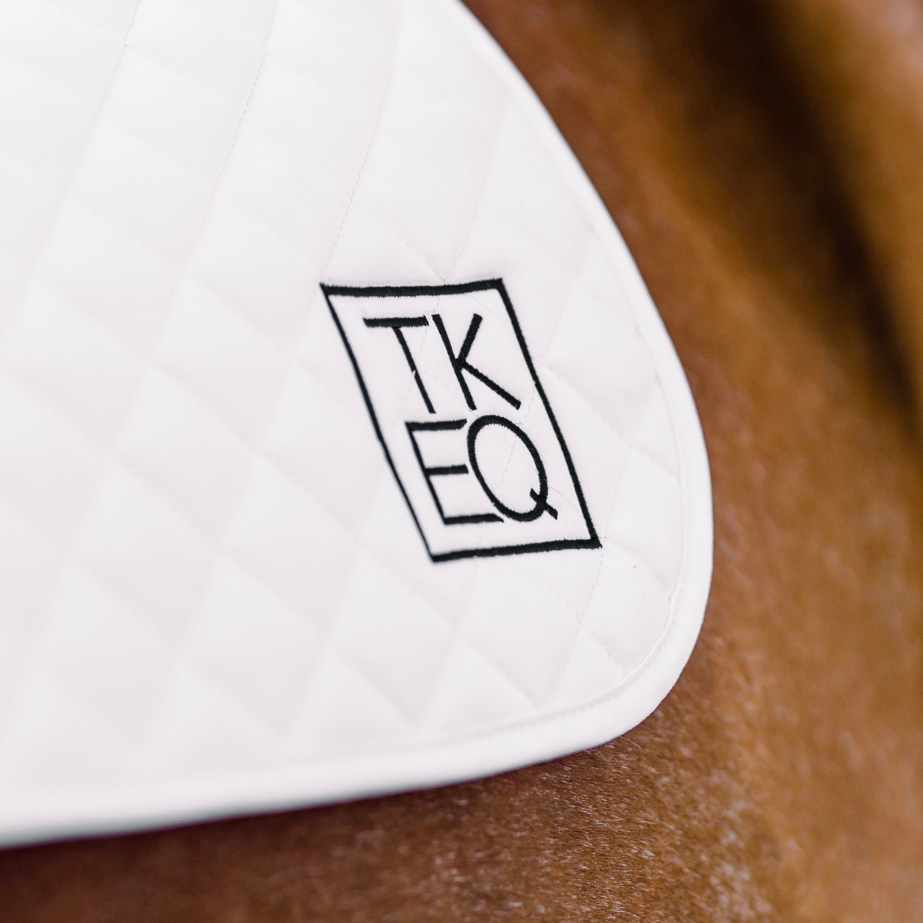 Close-up view of the TKEQ logo on the White TKEQ x SPR Jump Saddle Pad, as worn by a horse