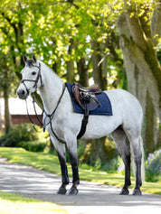 Side view of a gray horse wearing the Navy LeMieux Pillow Pad under a brown saddle while standing on a paved path outside