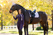 Front view of a female model wearing the Juniper LeMieux Philippa Mesh Base Layer while standing in front of a horse outside