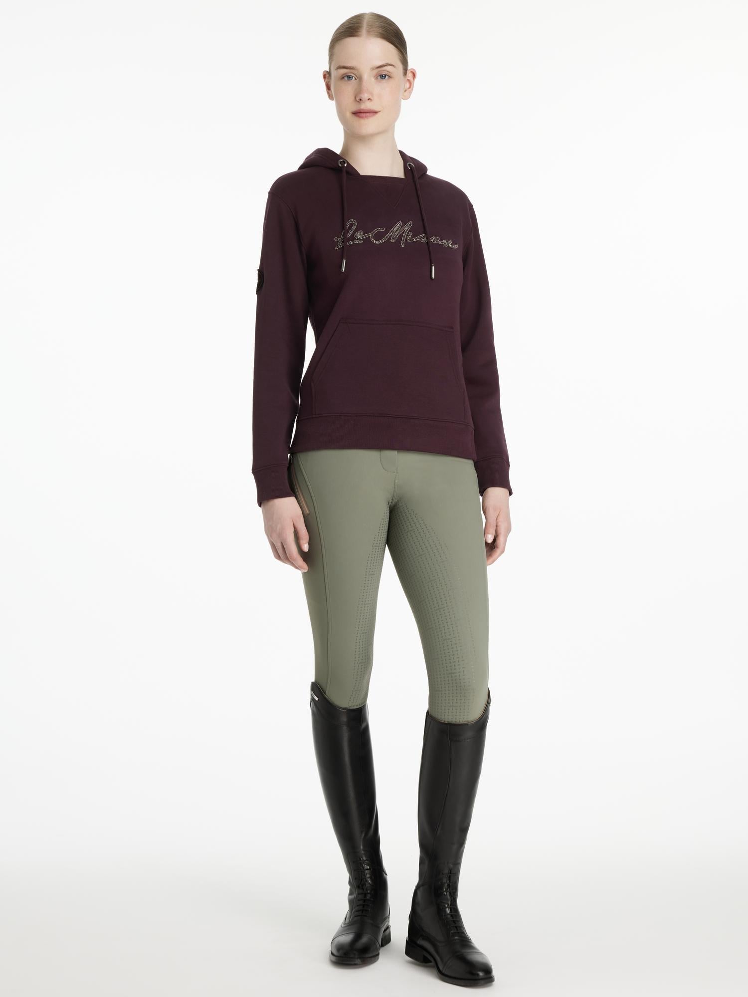Full body front view of a female model wearing the Damson LeMieux Elspeth Hoodie with green breeches on a plain background