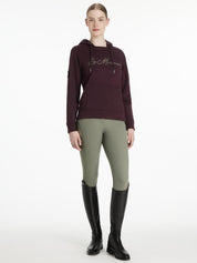 Full body front view of a female model wearing the Damson LeMieux Elspeth Hoodie with green breeches on a plain background