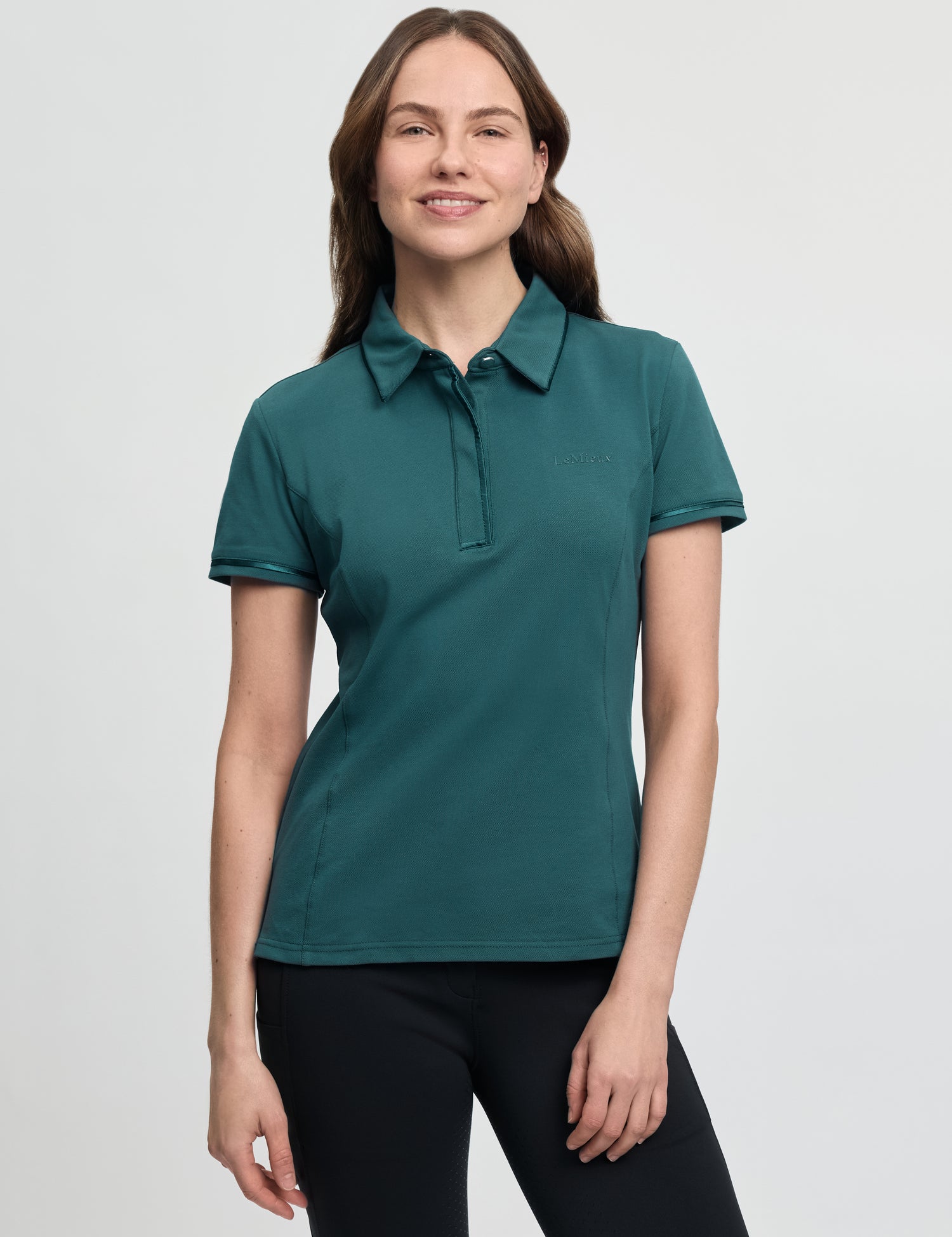 LeMieux Pixie polo shirt in jungle green with short sleeves, pointed collar, and partial button placket, shown on a model wearing black tights against a light gray background.
