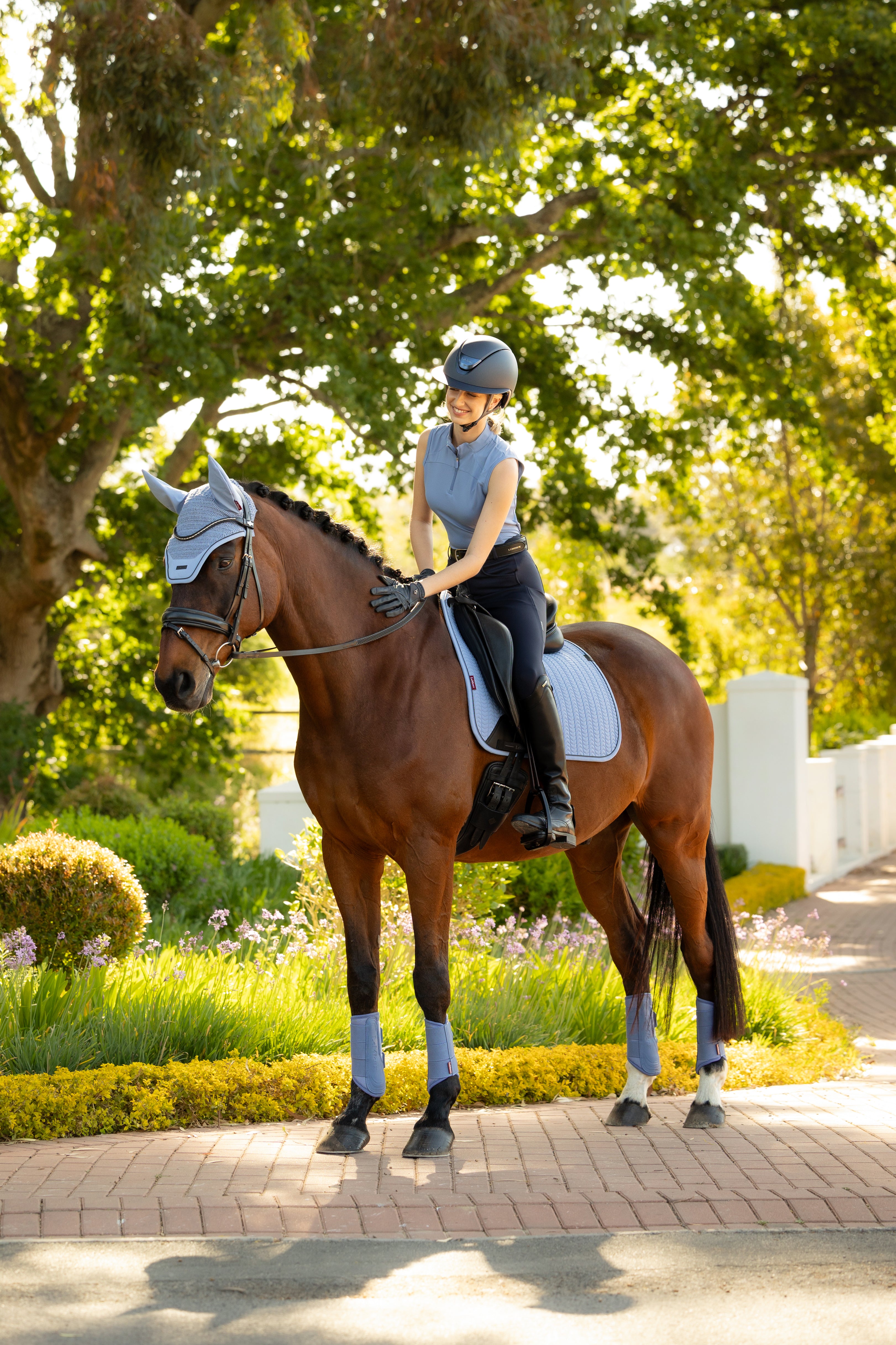 LeMieux Airflow sleeveless base layer in powder blue with quarter-zip high collar, worn with navy riding tights, black gloves, and tall black boots while sitting on a saddled horse outdoors.