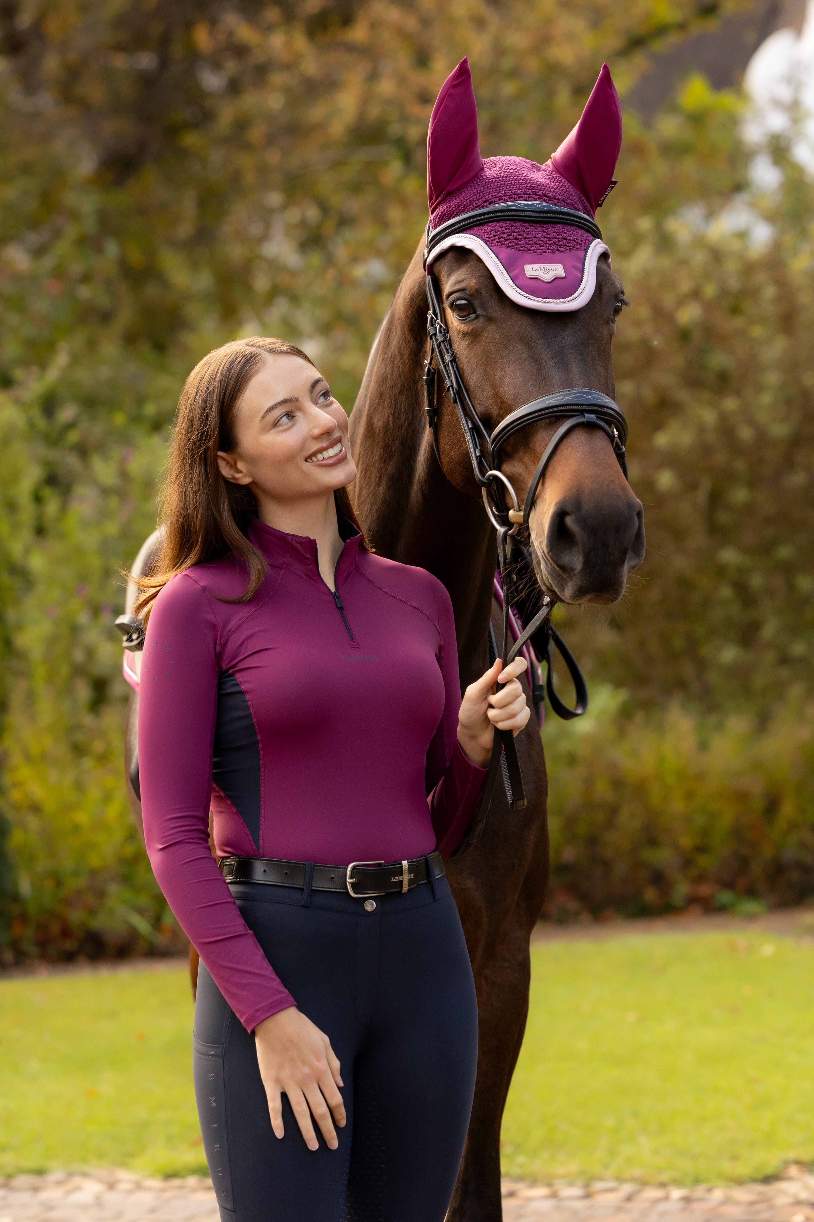 Front view of a model standing outside in front of a horse that is wearing the Blackberry LeMieux Loire Fly Hood
