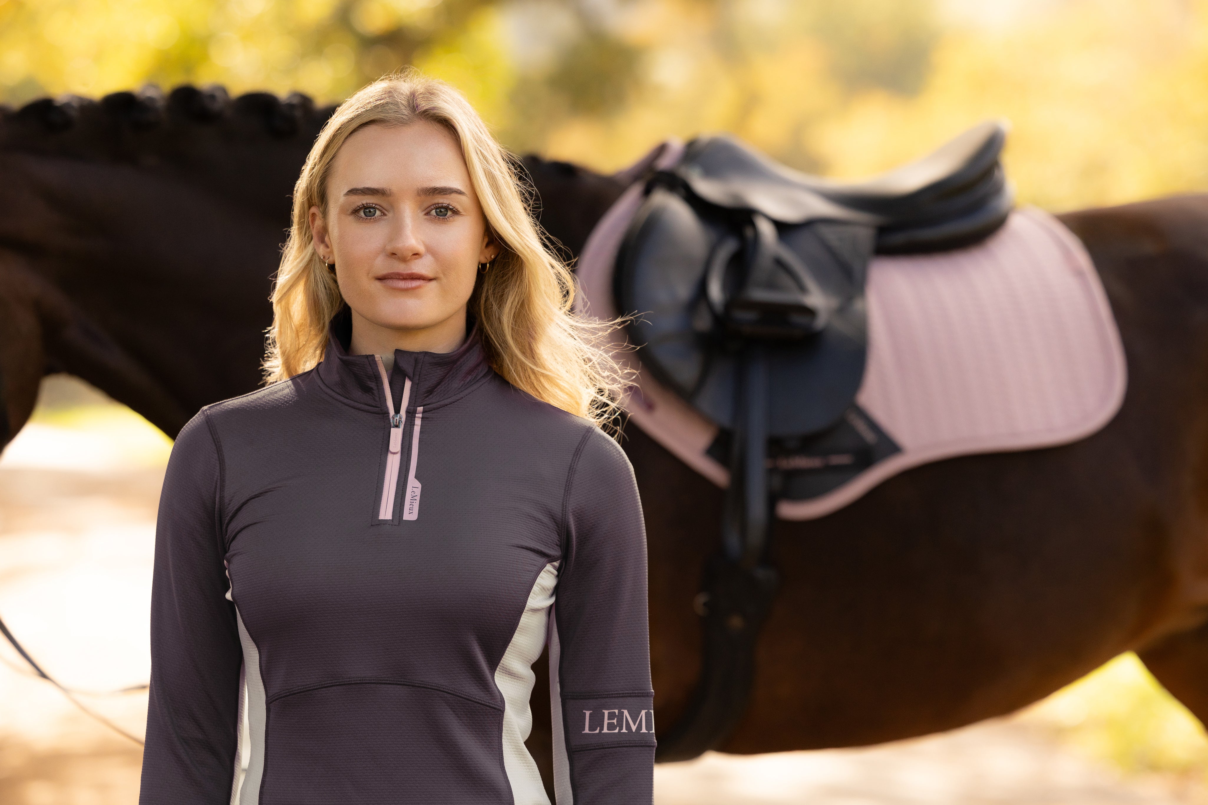 Front view of a female model wearing the Cocoa LeMieux Matilda Mid Layer while standing in front of a horse outside