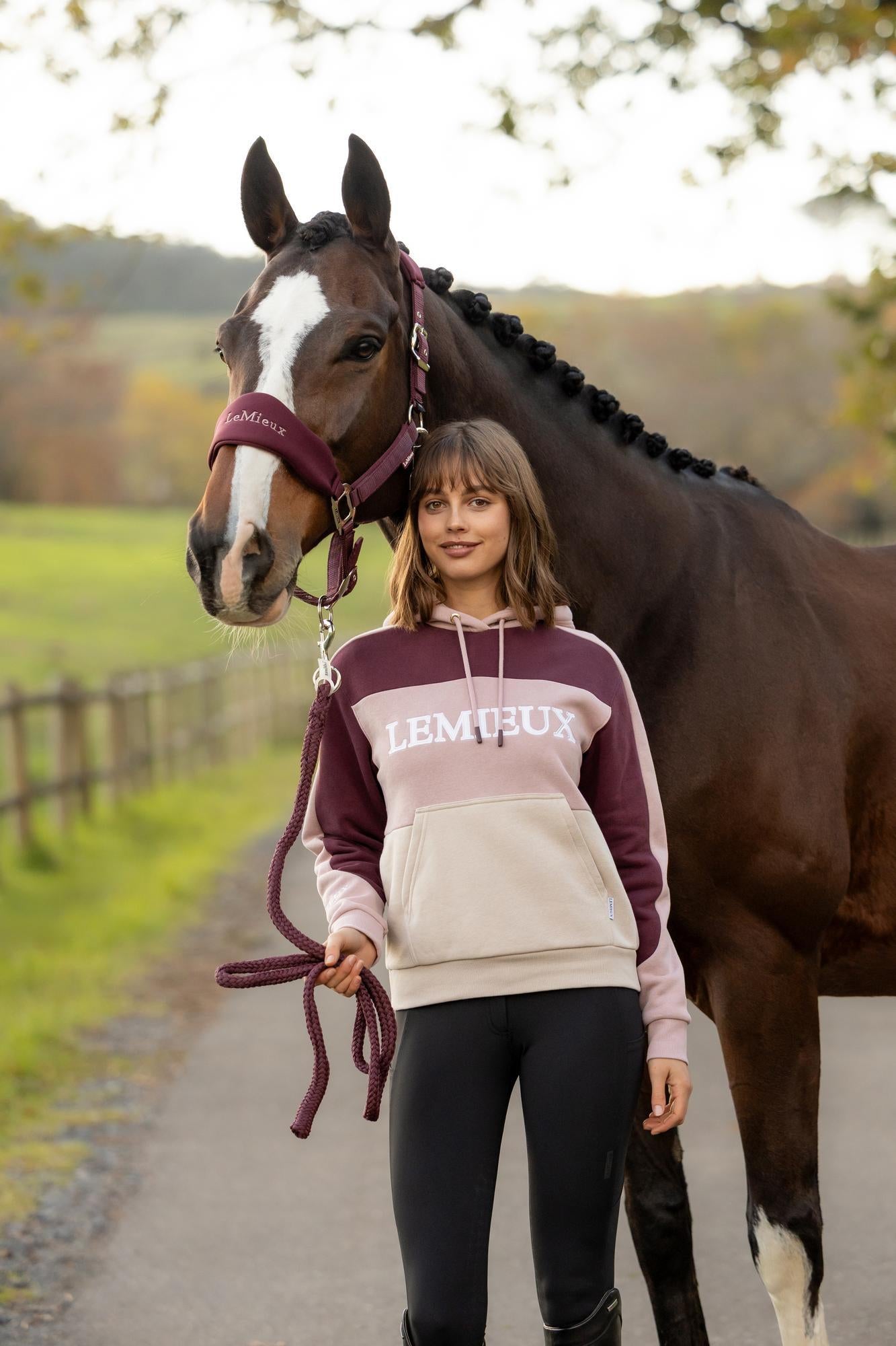 Front view of a female model wearing the Wild Rose LeMieux Kadie Color Block Hoodie while standing in front of a horse on a path outside