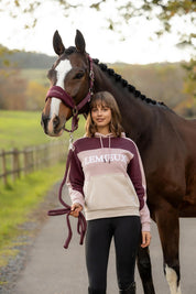 Front view of a female model wearing the Wild Rose LeMieux Kadie Color Block Hoodie while standing in front of a horse on a path outside