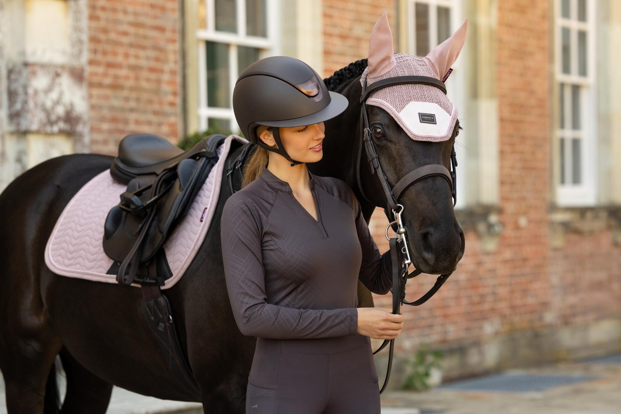 View of a female model wearing the Cocoa LeMieux Heritage Check Long Sleeve Base Layer while standing in front of a black horse