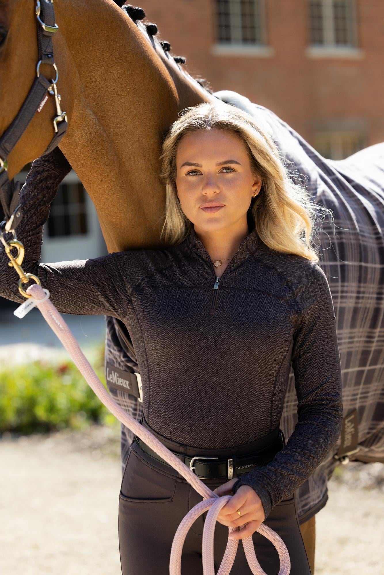 View of a female model wearing the Cocoa LeMieux Heritage Herringbone Base Layer while standing outside in front of a bay horse