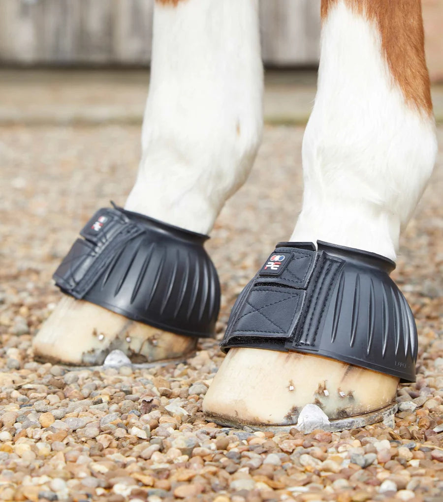 Vie wof a horse wearing the Black Premier Equine Rubber Bell Over Reach Boots while standing on a gravel path