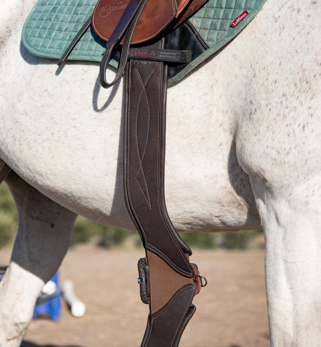 View of the Brown Total Saddle Fit StretchTec Shoulder Relief Girth – Jump hanging from a saddle that is worn by a gray horse