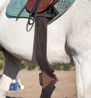 View of the Brown Total Saddle Fit StretchTec Shoulder Relief Girth – Jump hanging from a saddle that is worn by a gray horse