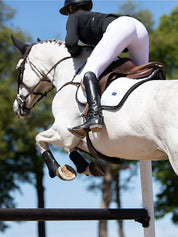 Angled back view of a female model riding a grey horse over a jump while the horse is wearing the Black Edition Equestrian Stockholm Modern White Jump Pad