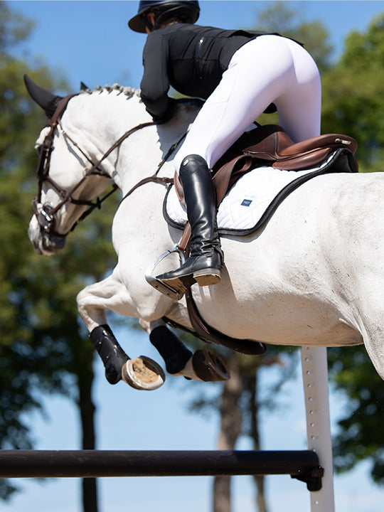 Angled back view of a female model riding a grey horse over a jump while the horse is wearing the Black Edition Equestrian Stockholm Modern White Jump Pad
