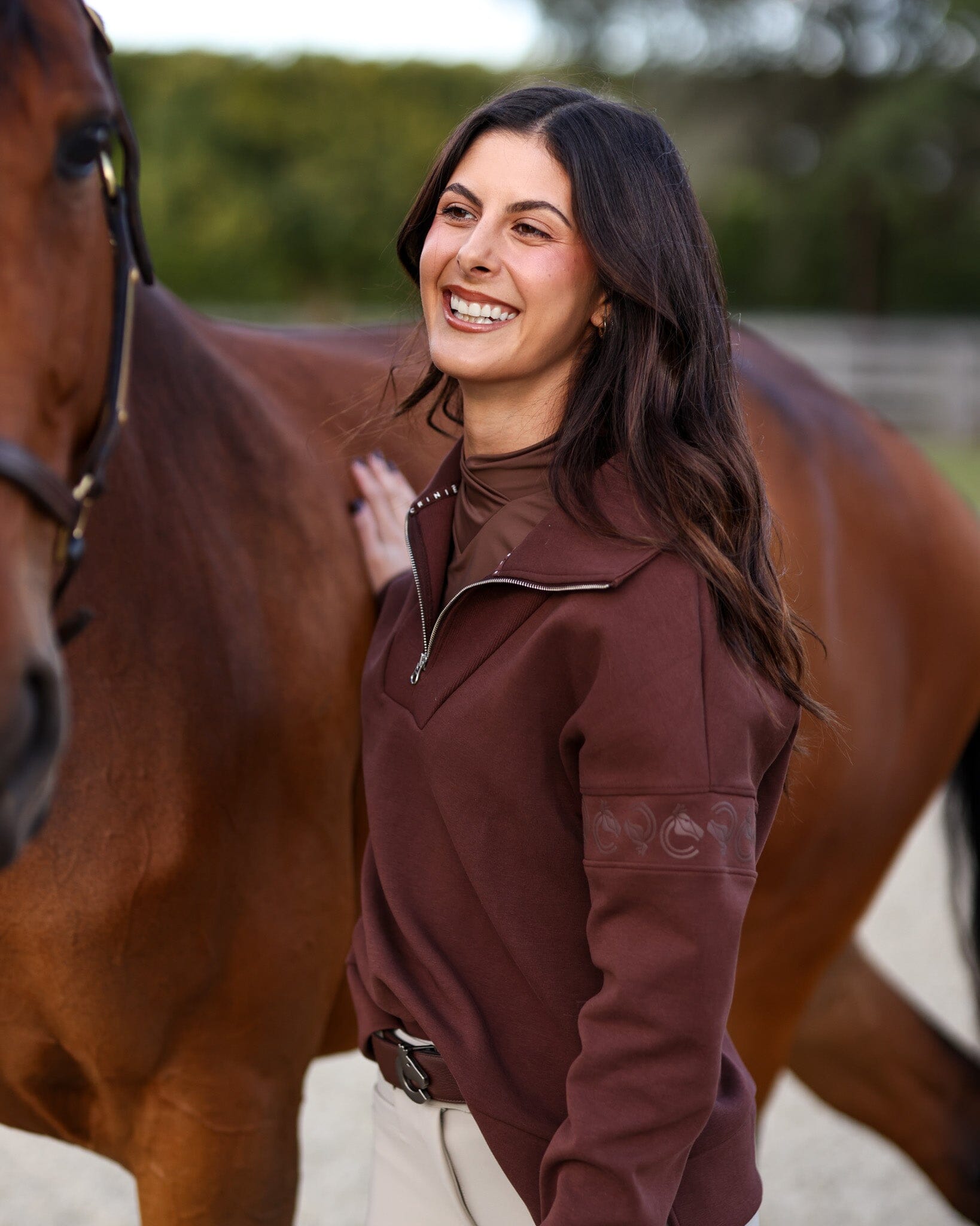 Angled front view of a female model wearing the Mocha Criniere Claire Sweater while standing next to a horse in an outdoor arena