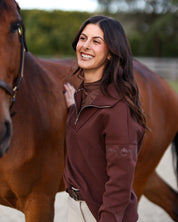 Angled front view of a female model wearing the Mocha Criniere Claire Sweater while standing next to a horse in an outdoor arena